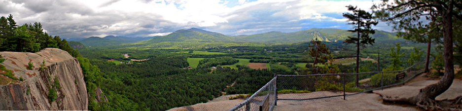 [Panoramic view of the heavily treed mountains and valley with many clouds in the sky. In the foreground is the fenced edge of the cliff.]